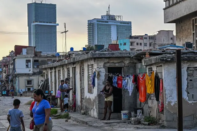 Varias personas hacen su vida diaria en un barrio de bajos recursos en La Habana. De fondo se ven edificios altos.